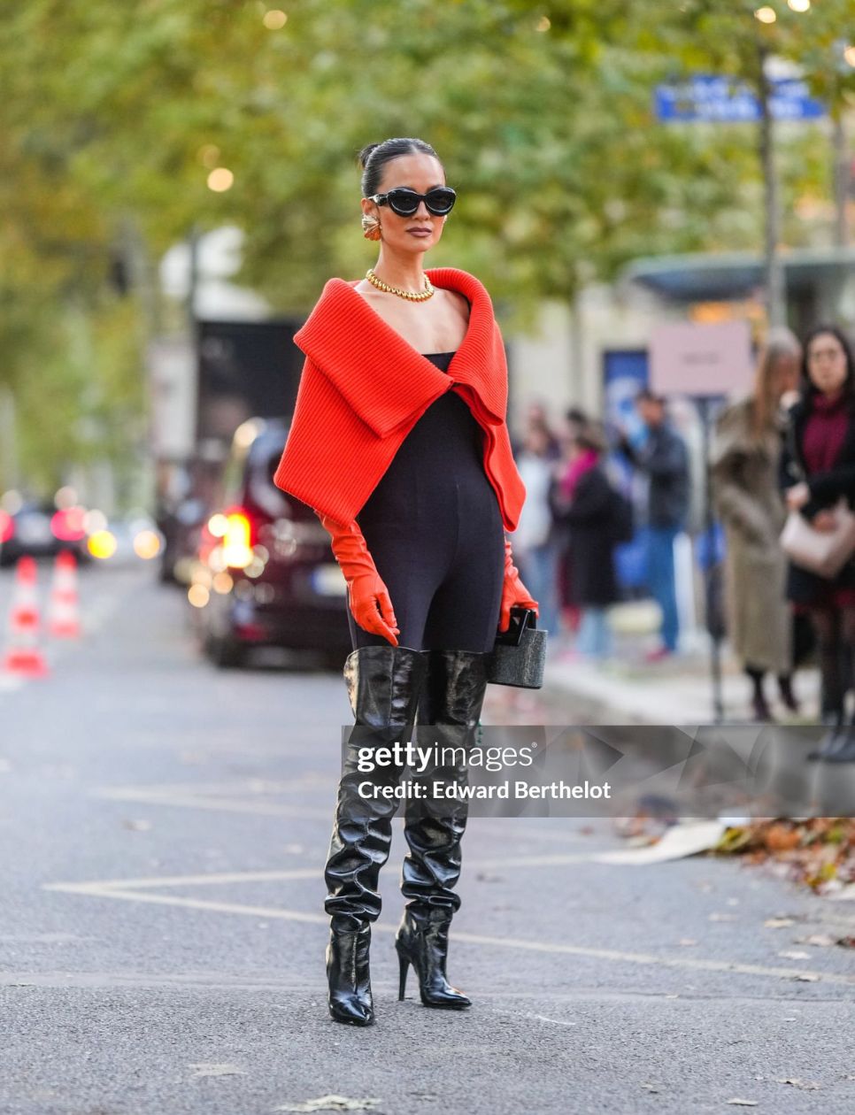 Anna Rosa Vitiello wears black puffy sunglasses from Loewe, gold large earrings, a gold large necklace, a red ribbed wool V-neck / oversized cropped cloak, red shiny leather high gloves, a black shoulder-off legging jumpsuit, a black embroidered rhinestones handbag, black shiny varnished leather heels pointed high boots / high waders , outside Akris, during Paris Fashion Week - Womenswear Spring/Summer 2023, on October 01, 2022 in Paris, France. (Photo by Edward Berthelot/Getty Images)