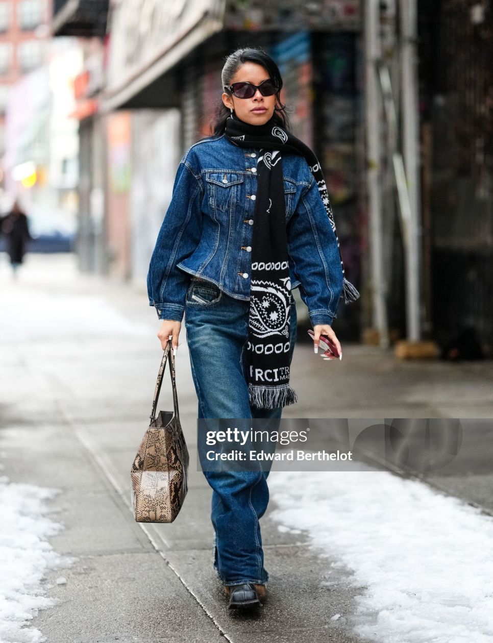 A guest wears black sunglasses, silver earrings, black white pattern/print scarf, navy blue buttoned up tailored denim jean jacket, light gray python snake skin pattern/print leather bag, navy blue washed loose denim jean pants, shiny black boots leather shoes, outside Monse, during the New York Fashion week Fall/Winter 2025 on February 9, 2025 in New York, New York. (Photo by Edward Berthelot/Getty Images)