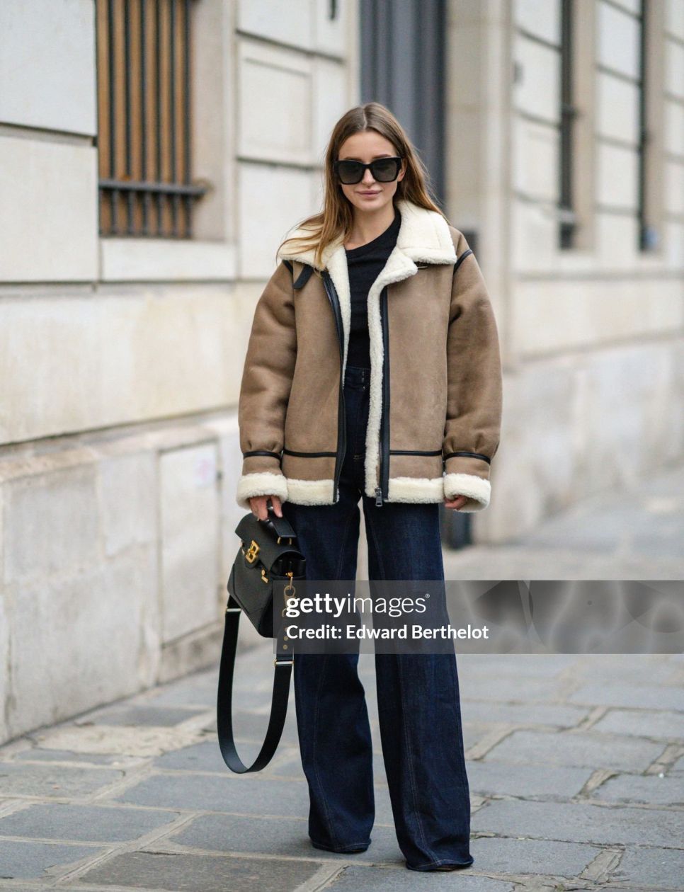 Diane Batoukina wears black sunglasses, a black t-shirt, a brown leather with black strap and white sheep interior oversized aviator coat, high waist navy blue denim palazzo / flared pants, a black shiny leather handbag from Balanciaga, black leather pumps heels shoes, during a street style fashion photo session, on December 15, 2021 in Paris, France. (Photo by Edward Berthelot/Getty Images) 