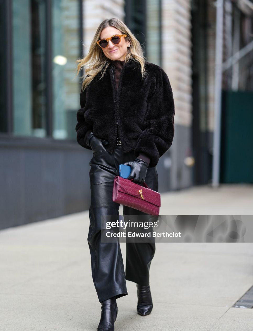 Guest wears light brown sunglasses, deep dark brown cropped faux fur jacket, shiny black leather gloves, shiny black straight loose leather pants, shiny black pointed toe heeled boots leather shoes, shiny burgundy bordeaux woven Bottega Veneta Andiamo clutch leather bag, outside Ulla Johnson, during the New York Fashion week Fall/Winter 2025 on February 9, 2025 in New York, New York. (Photo by Edward Berthelot/Getty Images)