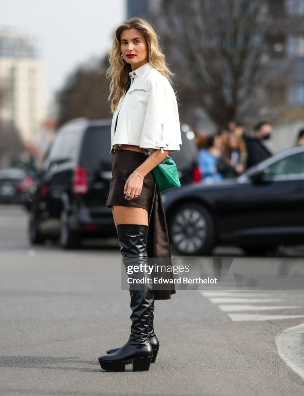 Xenia Adonts wears a white leather cropped cloak jacket, a dark brown satin tube short skirt with a long back-train, a green shiny leather with large embossed logo pattern crossbody bag from Prada, a black shoulder-off / cropped top from Prada, a silver chain bracelet, gold rings, black shiny leather platform soles / block heels waders / high boots from Prada, outside the Prada fashion show, during the Milan Fashion Week Fall/Winter 2022/2023 on February 24, 2022 in Milan, Italy. (Photo by Edward Berthelot/Getty Images)