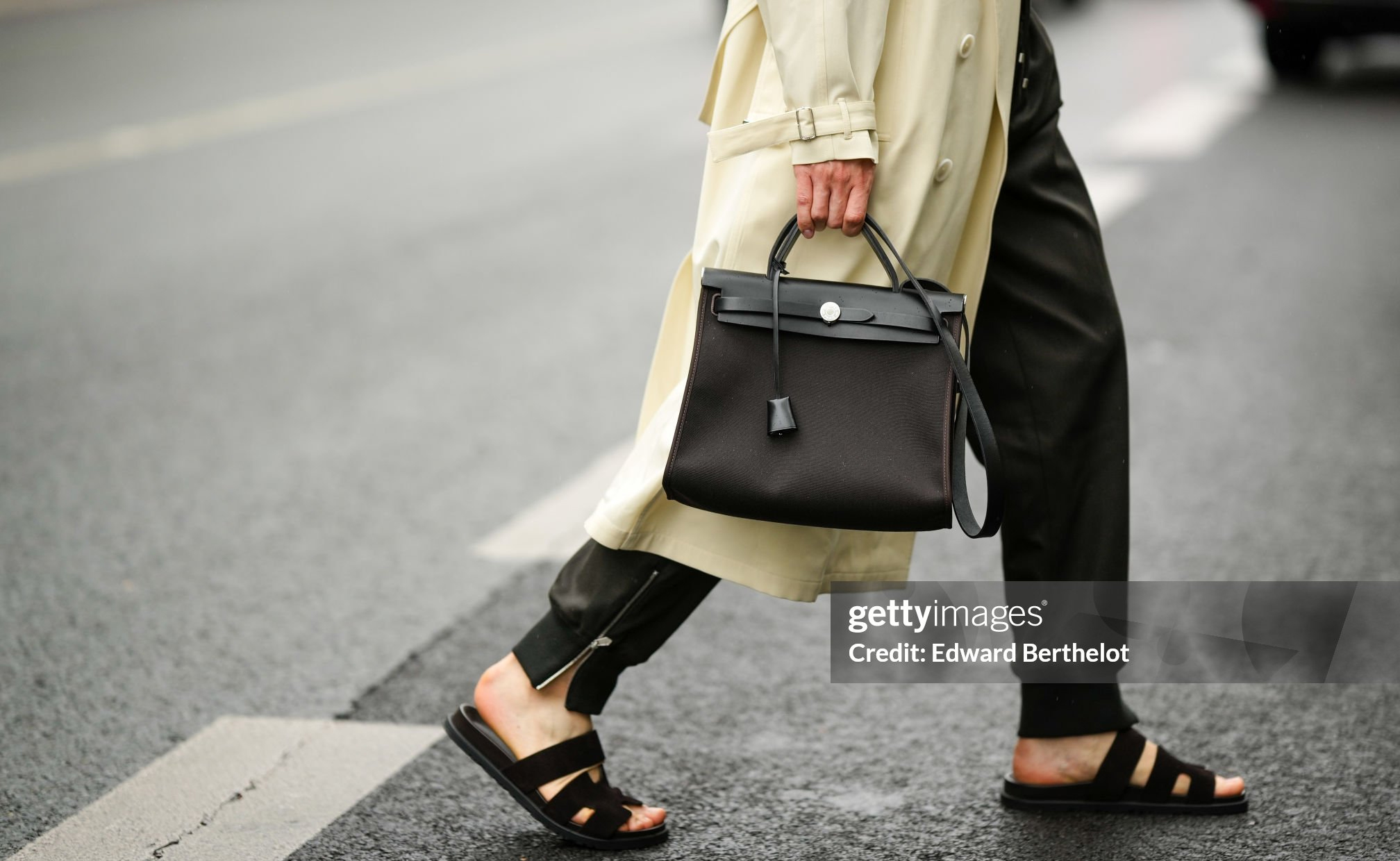 PARIS, FRANCE - JUNE 25: Lukas Osiecki wears a pale yellow long trench coat, black satin sport pants, a black shiny leather and fabric handbag from Hermès, black suede Chypre sandals from Hermès, outside the Hermès show, during Paris Fashion Week - Menswear Spring/Summer 2023, on June 25, 2022 in Paris, France. (Photo by Edward Berthelot/Getty Images)