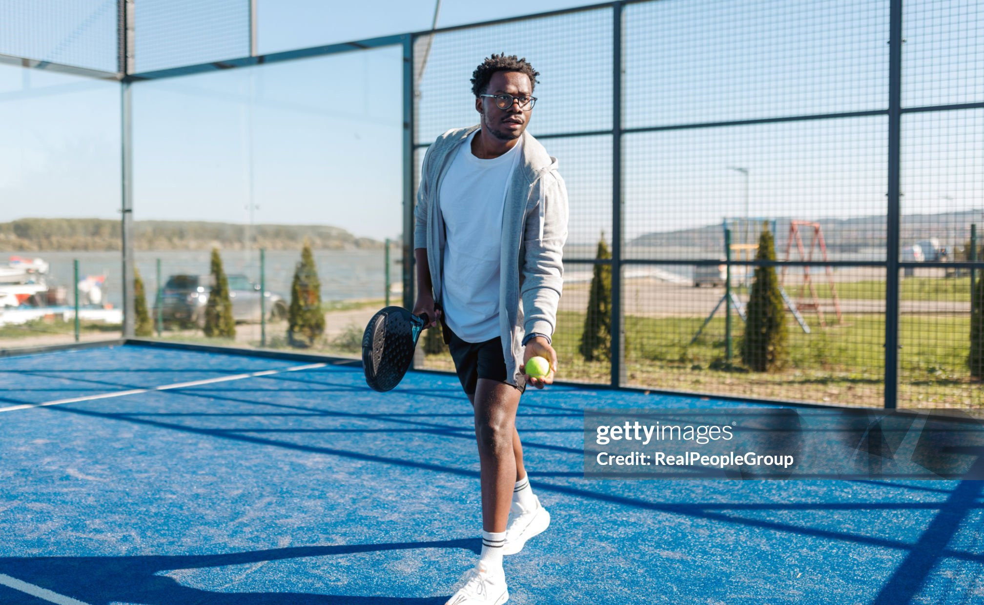 Young black man is about to serve a padel ball in a blue paddle tennis court near a river, he's holding a racket and a ball and looking intensely Getty Image