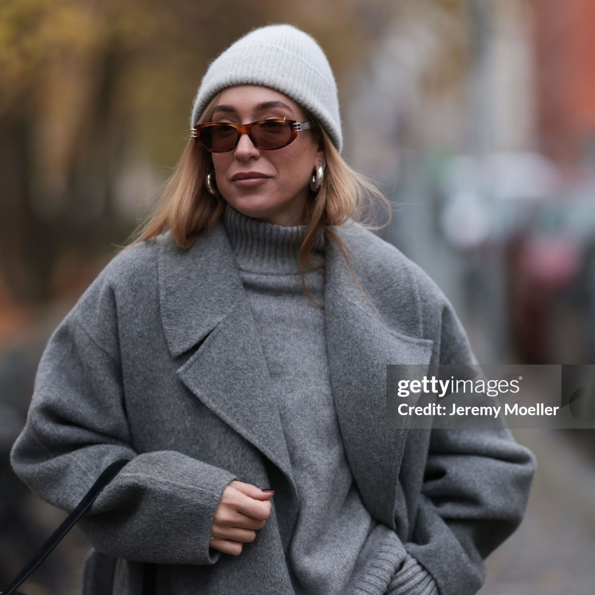 BERLIN, GERMANY - NOVEMBER 14: Sonia Lyson is seen wearing a light grey knit beanie from Le Bonnet; chunky silver hoop earrings; oval sunglasses with Havana frame and silver details from Givenchy; a calf-length grey wool coat with notch lapels from & Other Stories; a light grey knit sweater with ribbed turtleneck and cuffs from Sézane; and a rectangular black suede handbag from Elleme on November 14, 2024 in Berlin, Germany. (Photo by Jeremy Moeller/Getty Images)