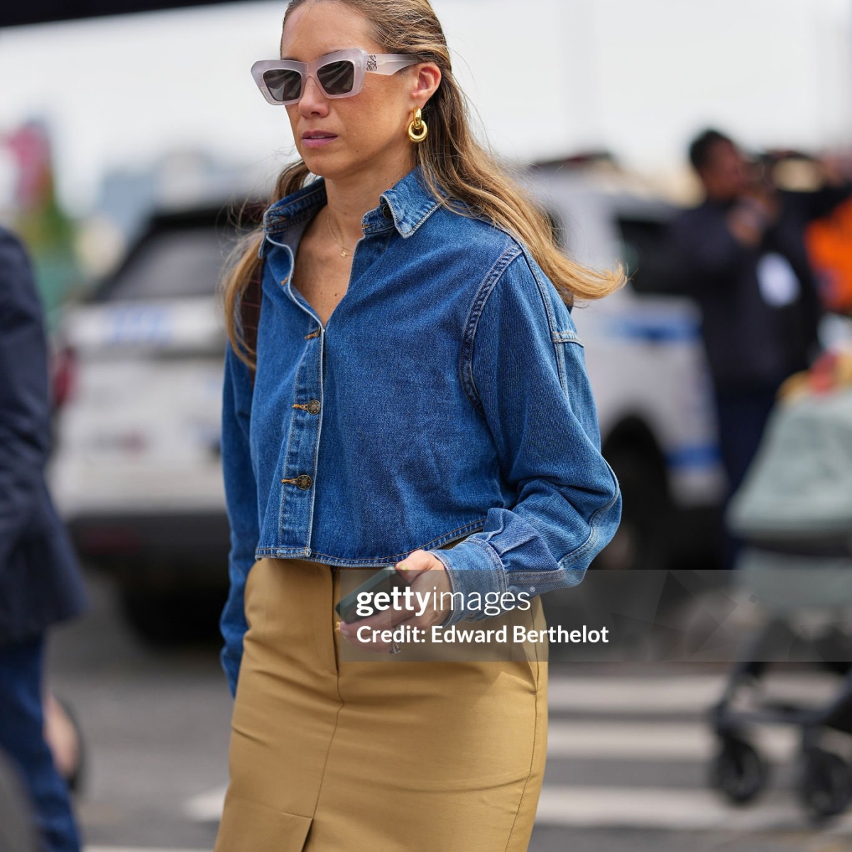 NEW YORK, NEW YORK - SEPTEMBER 15: A guest wears long light brown hair pulled back from the face with a center part, translucent pale pink oversized cat-eye plastic sunglasses, chunky gold hoop earrings and a thin gold chain necklace, a cropped blue denim jacket with pointed collar, long sleeves and button-front closure, a high-waisted tan cotton pencil skirt with a front center slit, outside Coach, during New York Fashion Week, on September 15, 2025 in New York, New York (Photo by Edward Berthelot/Getty Images)